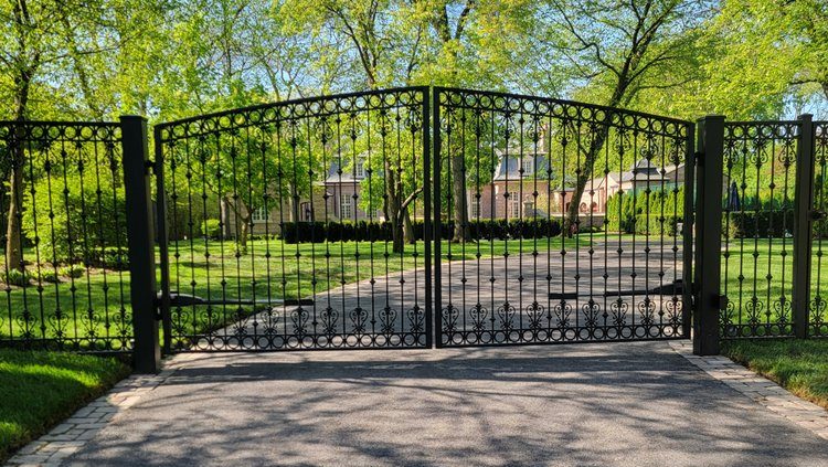 A wrought iron gate flanked by brick posts opens to a driveway lined with lush green trees. Sunlight filters through the leaves, creating a serene, inviting scene.