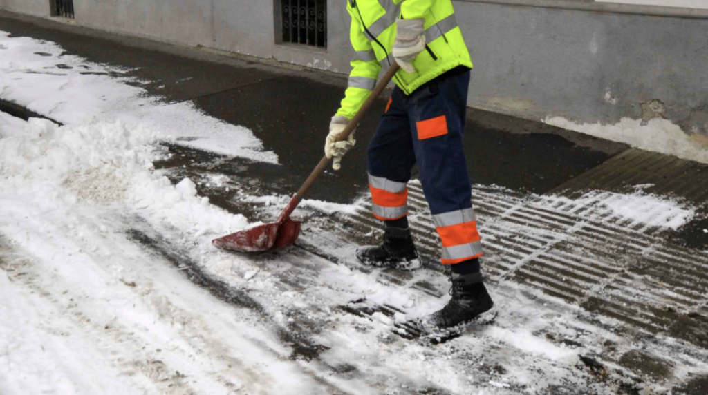 A person wearing a bright safety jacket and dark pants with orange stripes shovels snow off a sidewalk, revealing clear pathway, in an urban setting.